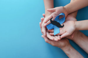 Family hands holding light blue ribbonwith mustache on blue background , Prostate Cancer Awareness, Men health awareness, November Blue